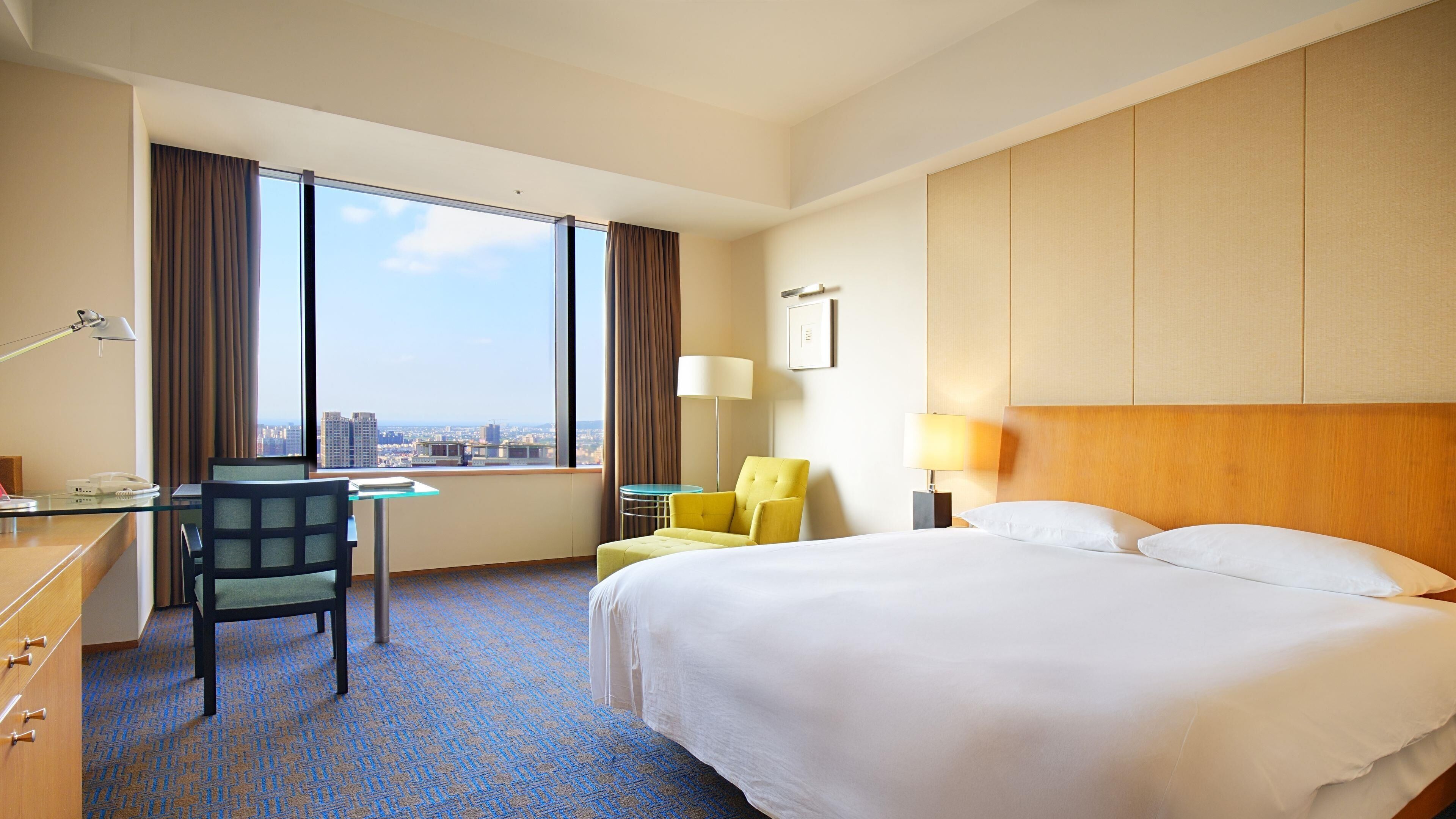 A hotel room with a large bed, a desk, a chair, a small table, and a view of the city through large windows.