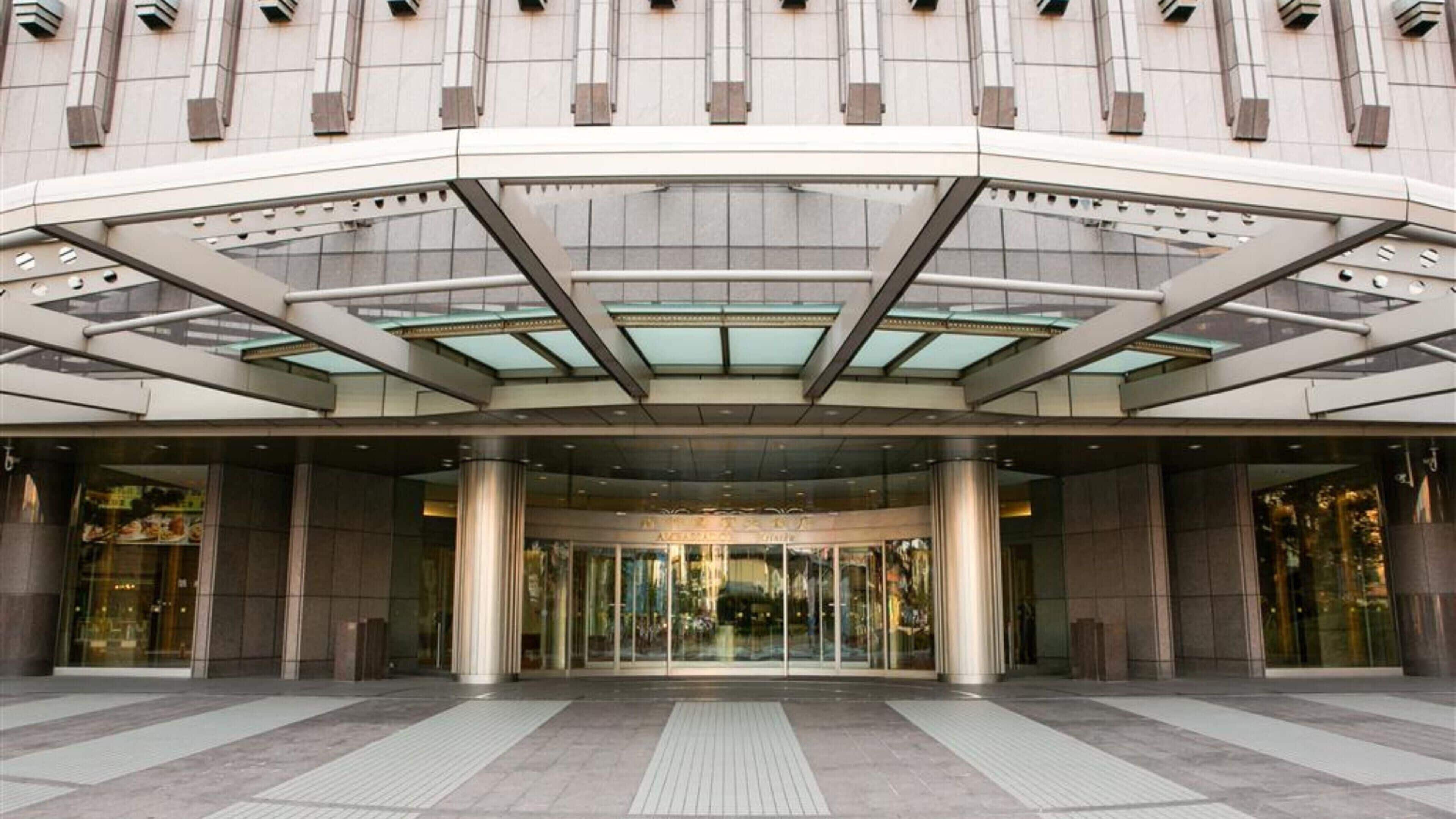 A modern hotel entrance with a glass canopy, large columns, and a tiled ground floor.