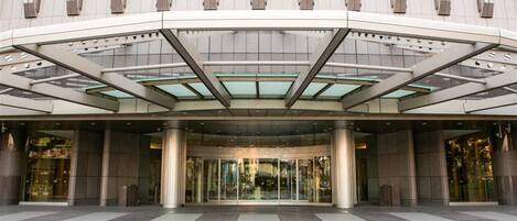 A modern hotel entrance with a glass canopy, large columns, and a tiled ground floor.