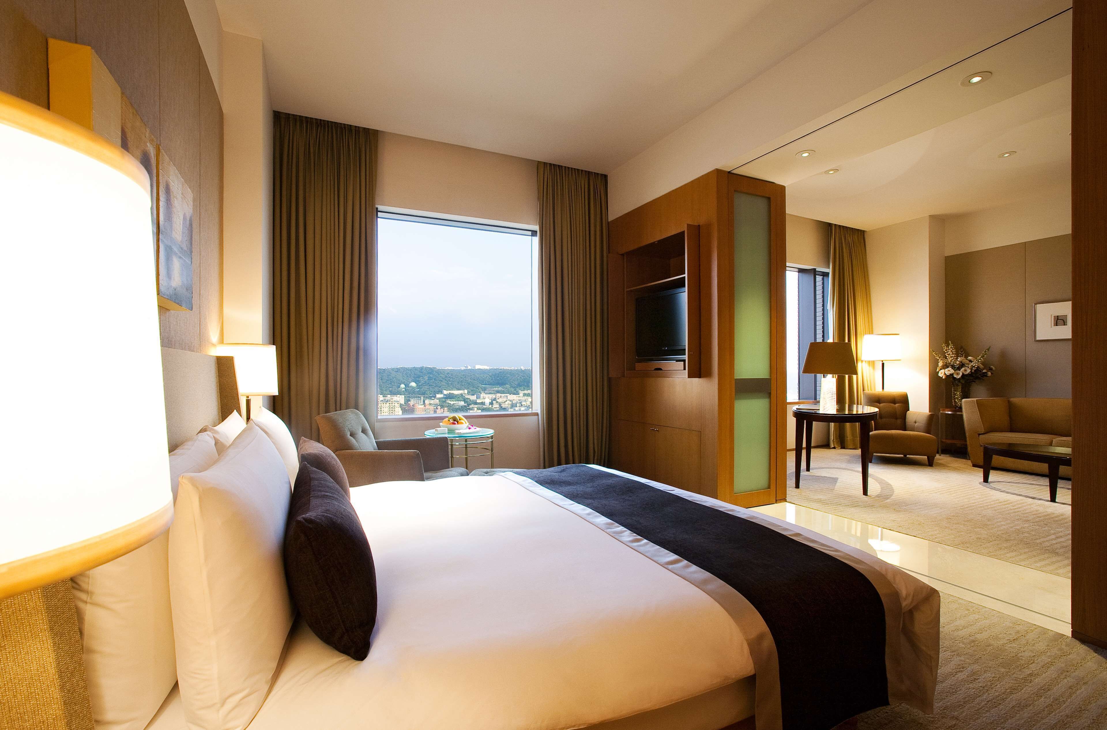 A hotel room with a large bed, a TV, a seating area with a sofa and chairs, a small table, and a view of the city through the window.