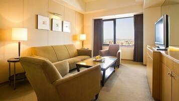 Habitación de hotel con sofá, dos sillones, una mesa de centro, televisor y vista a la ciudad desde la ventana.