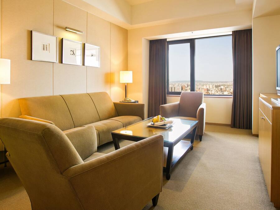 Habitación de hotel con sofá, dos sillones, una mesa de centro, televisor y vista a la ciudad desde la ventana.