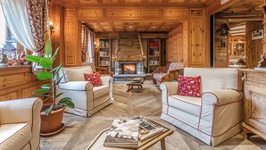 A cozy log cabin interior with a fireplace, plush seating, and a bookshelf.