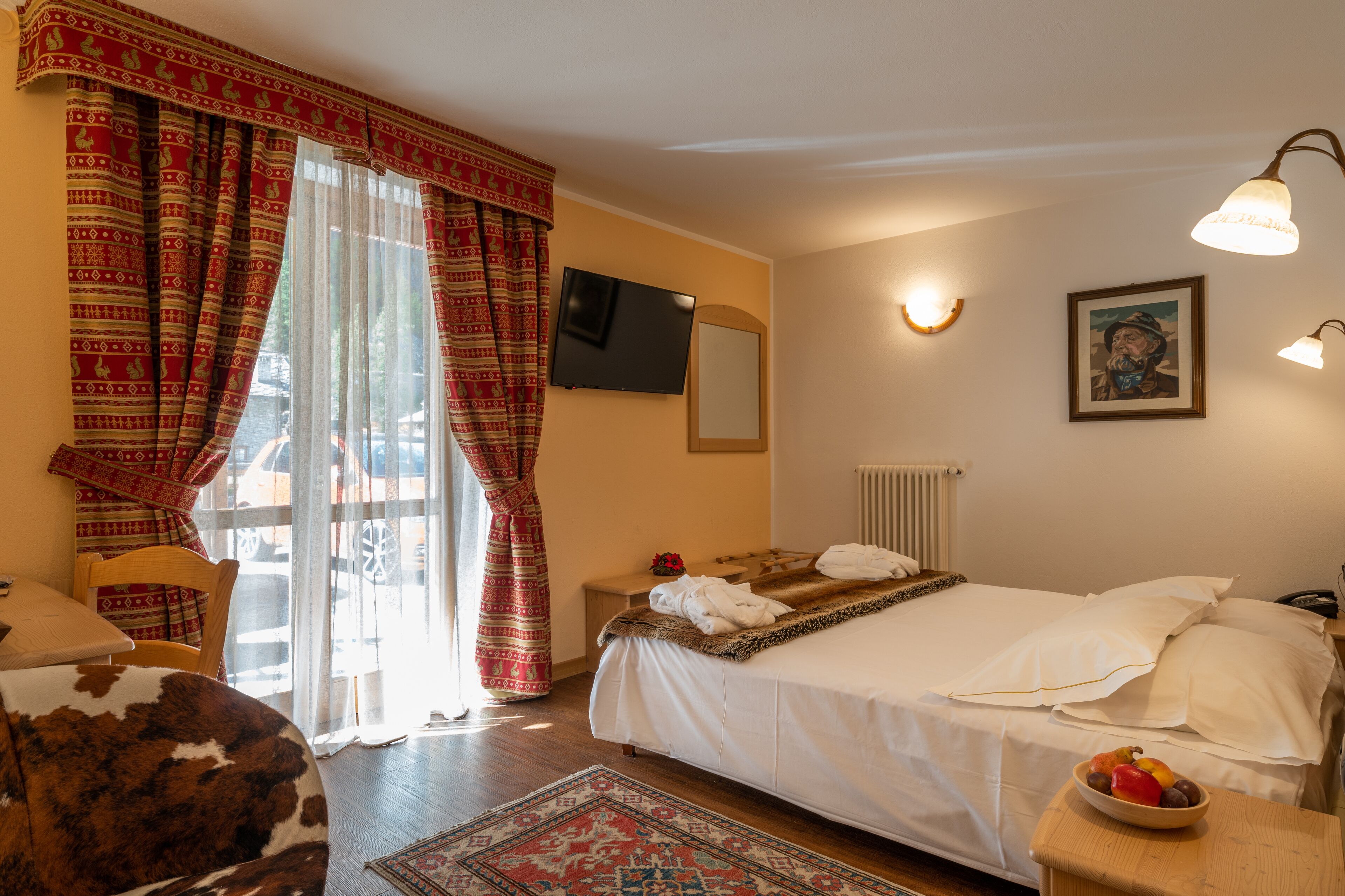 Ein Hotelzimmer mit einem Bett, einem Fernseher, einem Fenster mit Vorhängen, einem Nachttisch mit einer Obstschale und einem Holzstuhl.
