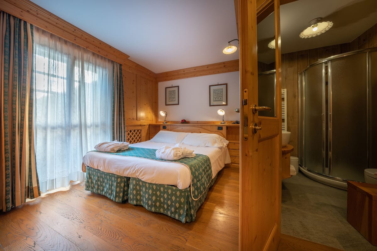 Een hotelkamer met een houten vloer, een bed met een bedspreide met een groen patroon, een groot raam met dunne gordijnen en een douchecel met een glazen deur.