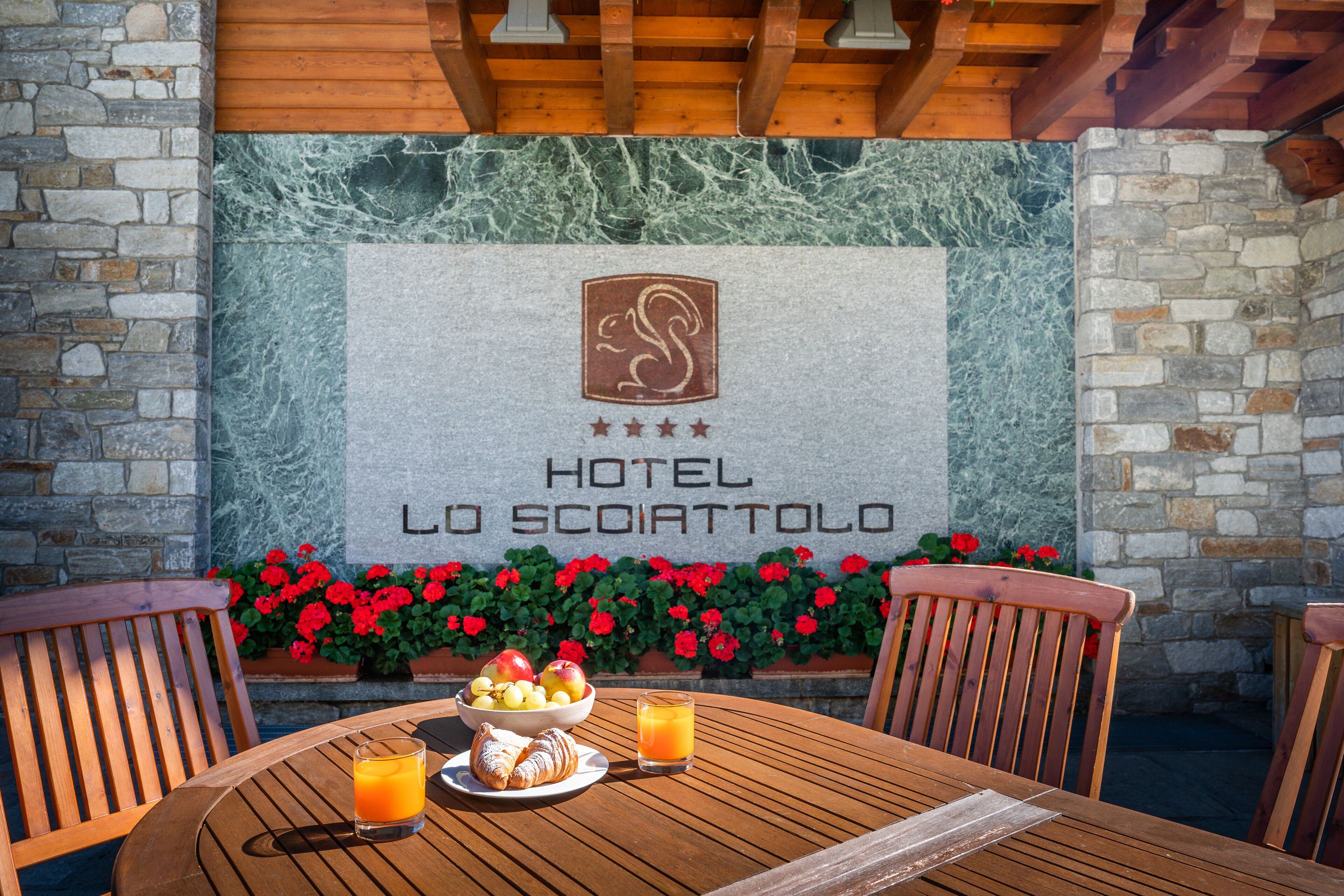 A wooden table with breakfast items, a stone wall with a hotel sign, and red flowers.