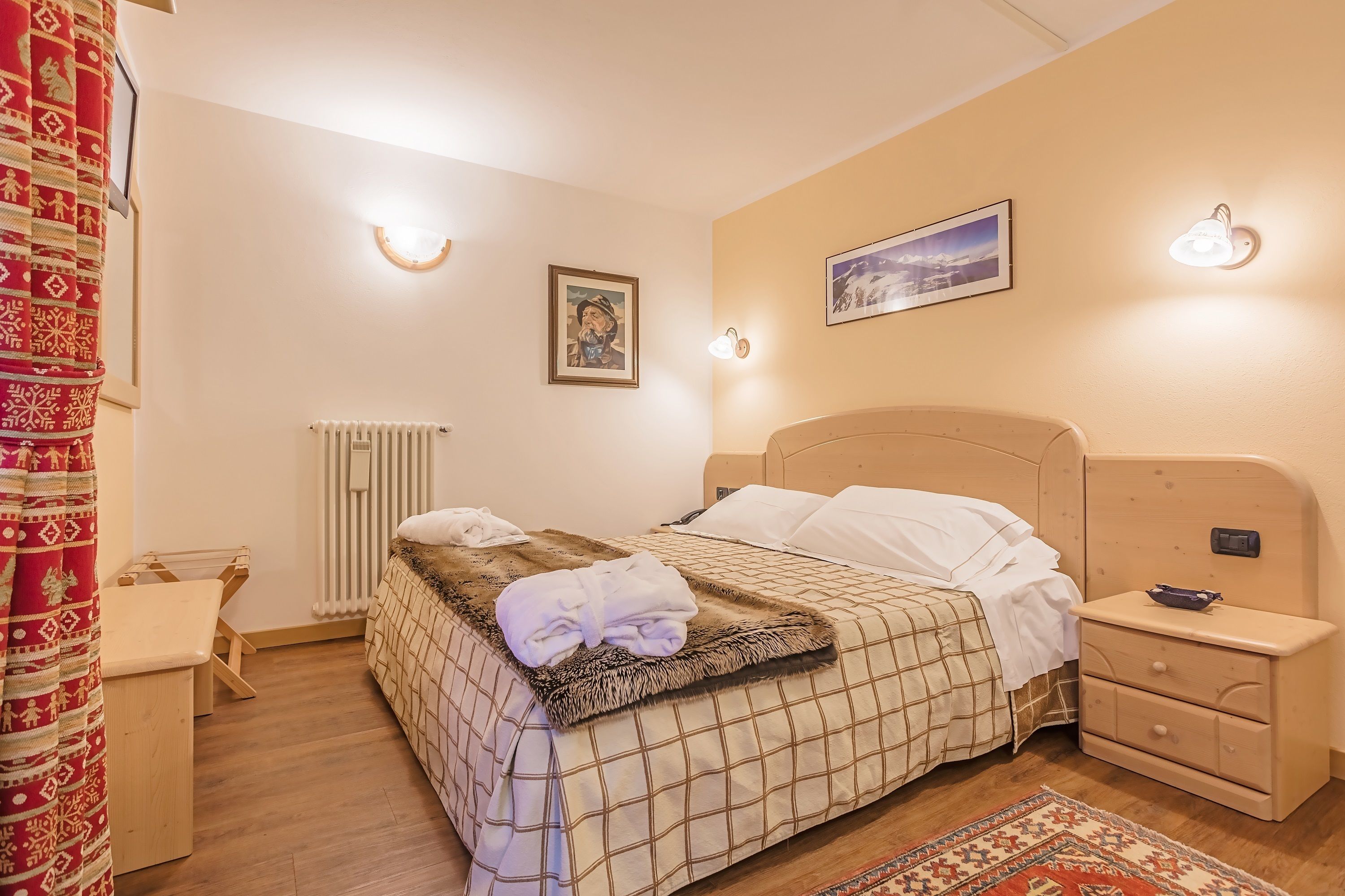 Un letto ben rifatto con copriletto a quadretti, una morbida coperta e una coppia di asciugamani piegati. C'è un comodino con una lampada e un tavolino con un cassetto. La stanza dispone di un termosifone, un quadro incorniciato a parete e un tappeto decorato sul pavimento.