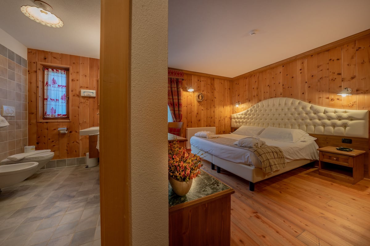 Een kamer met houten lambrisering, een bed, een badkamer met toilet en een raam met gordijn.