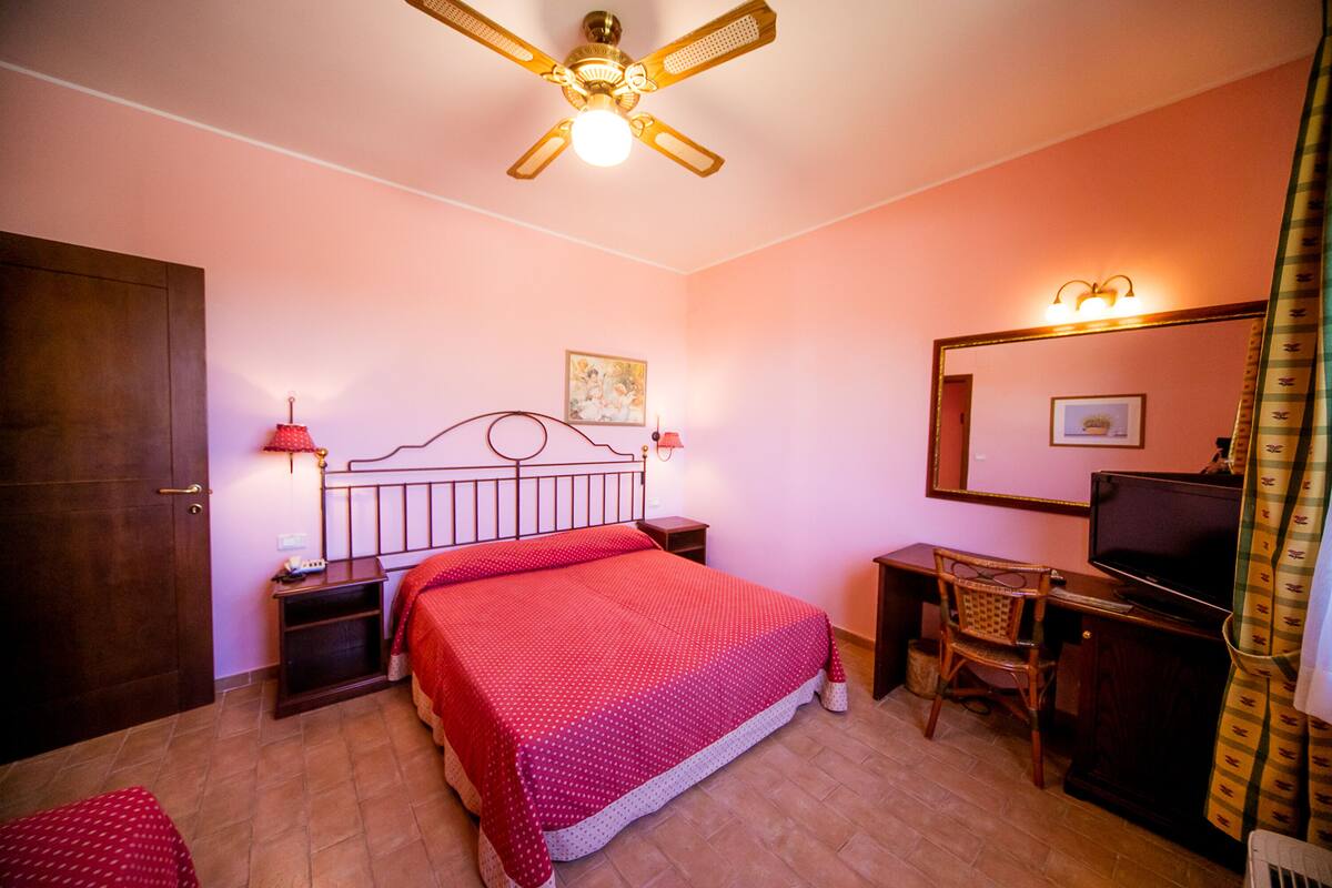 Een slaapkamer met een bed, een bureau, een stoel, een televisie, een plafondventilator en een deur.