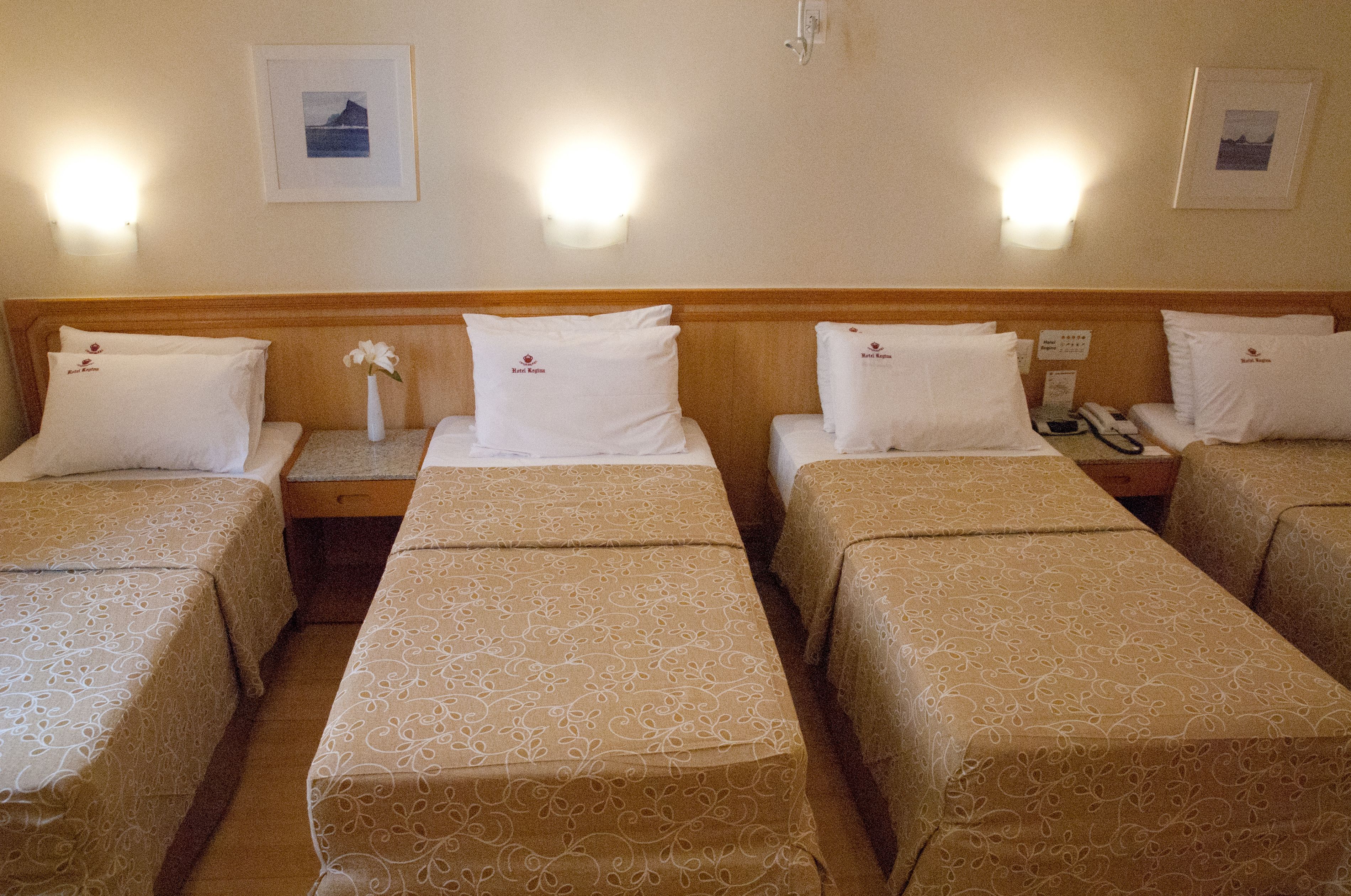 Quarto de hotel com quatro camas de solteiro, cada uma com cabeceira de madeira e roupa de cama branca. Há luminárias fixadas na parede e quadros emoldurados.