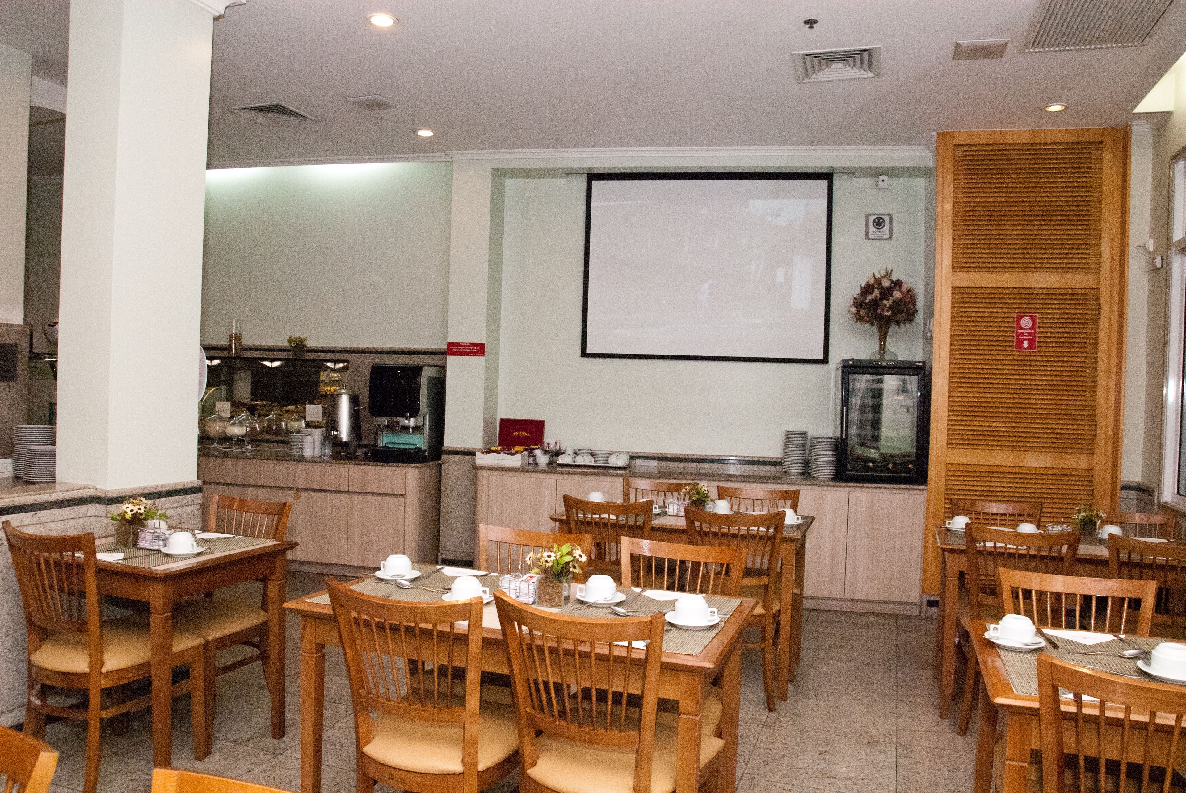 Un comedor con mesas y sillas de madera, una cocina al fondo y una pantalla de proyector en la pared.