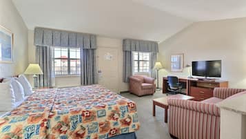 Una habitación de hotel con una cama, dos sillas, un escritorio con televisor y ventanas grandes con cortinas.