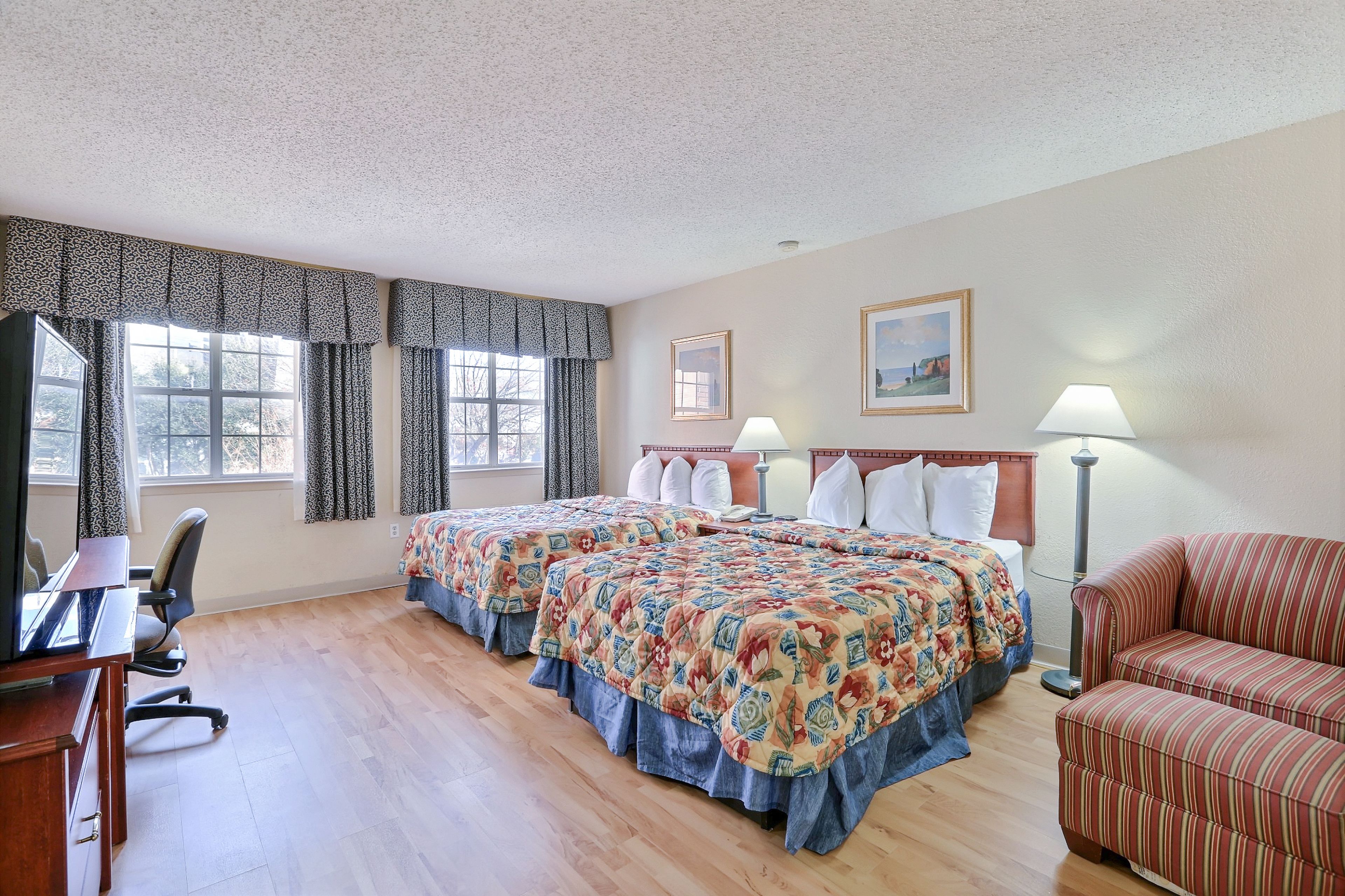 A hotel room with two beds, a desk, a chair, a sofa, and two lamps.