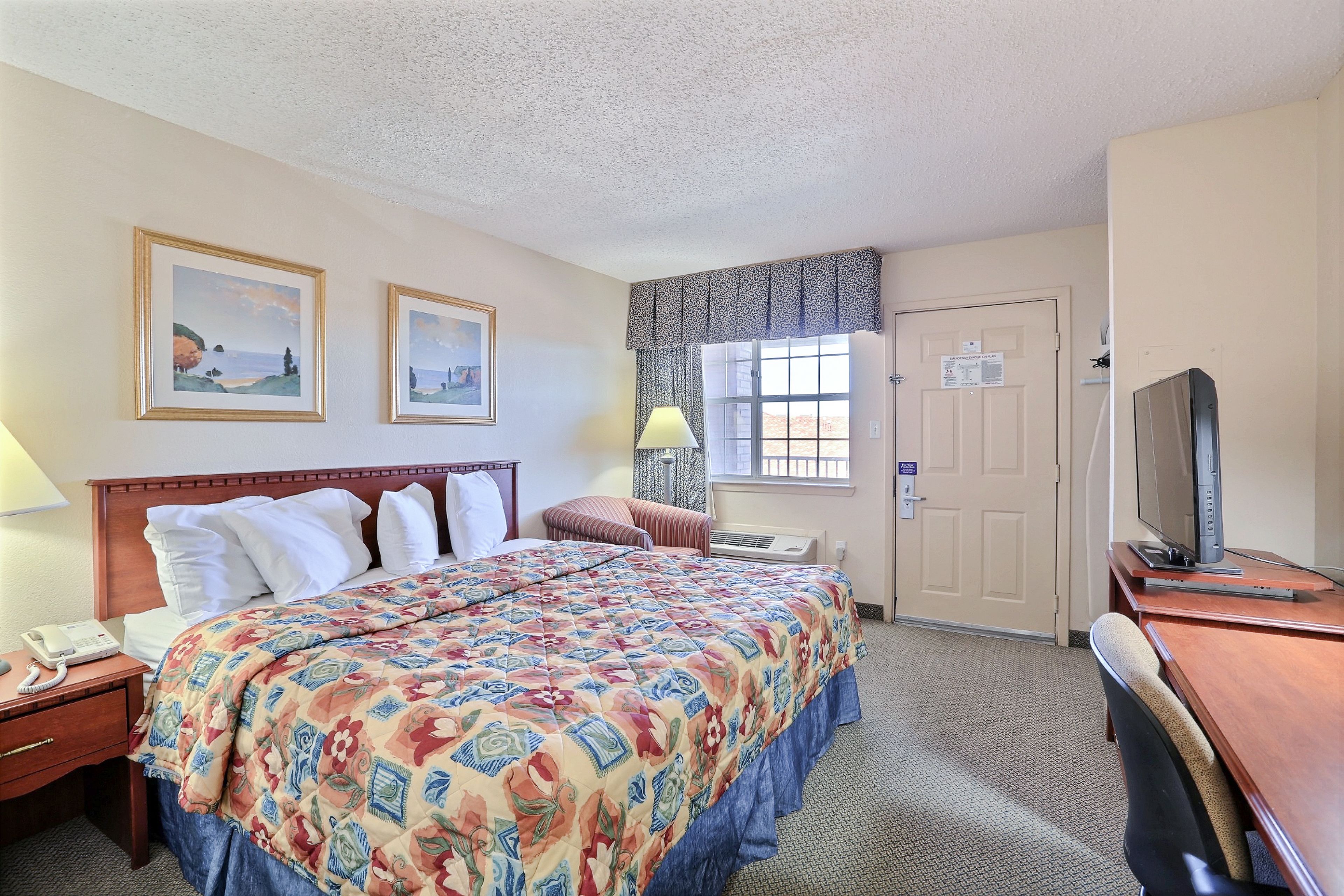 A hotel room with a large bed, a desk with a television, a window with curtains, and two framed pictures on the wall.