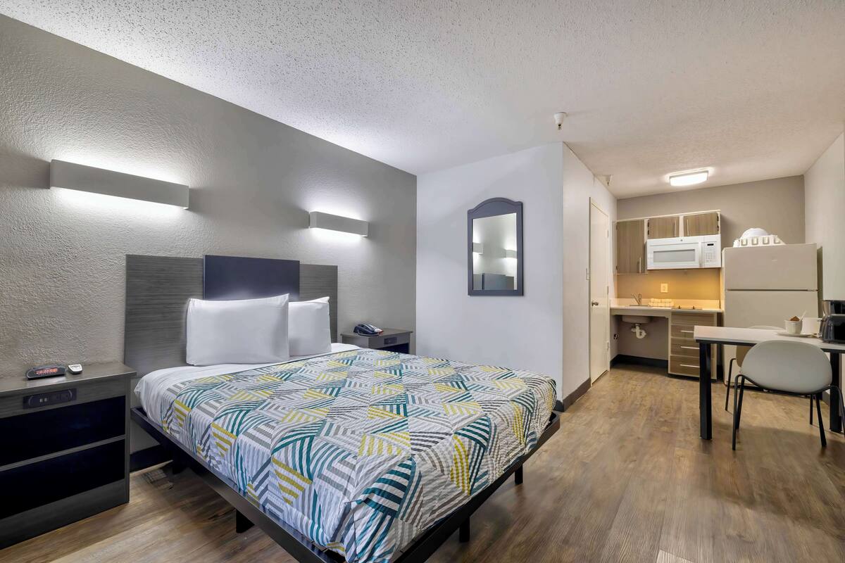 A modern hotel room with a bed, bedside table, and a small desk with a chair. There is a kitchenette with a microwave and refrigerator.