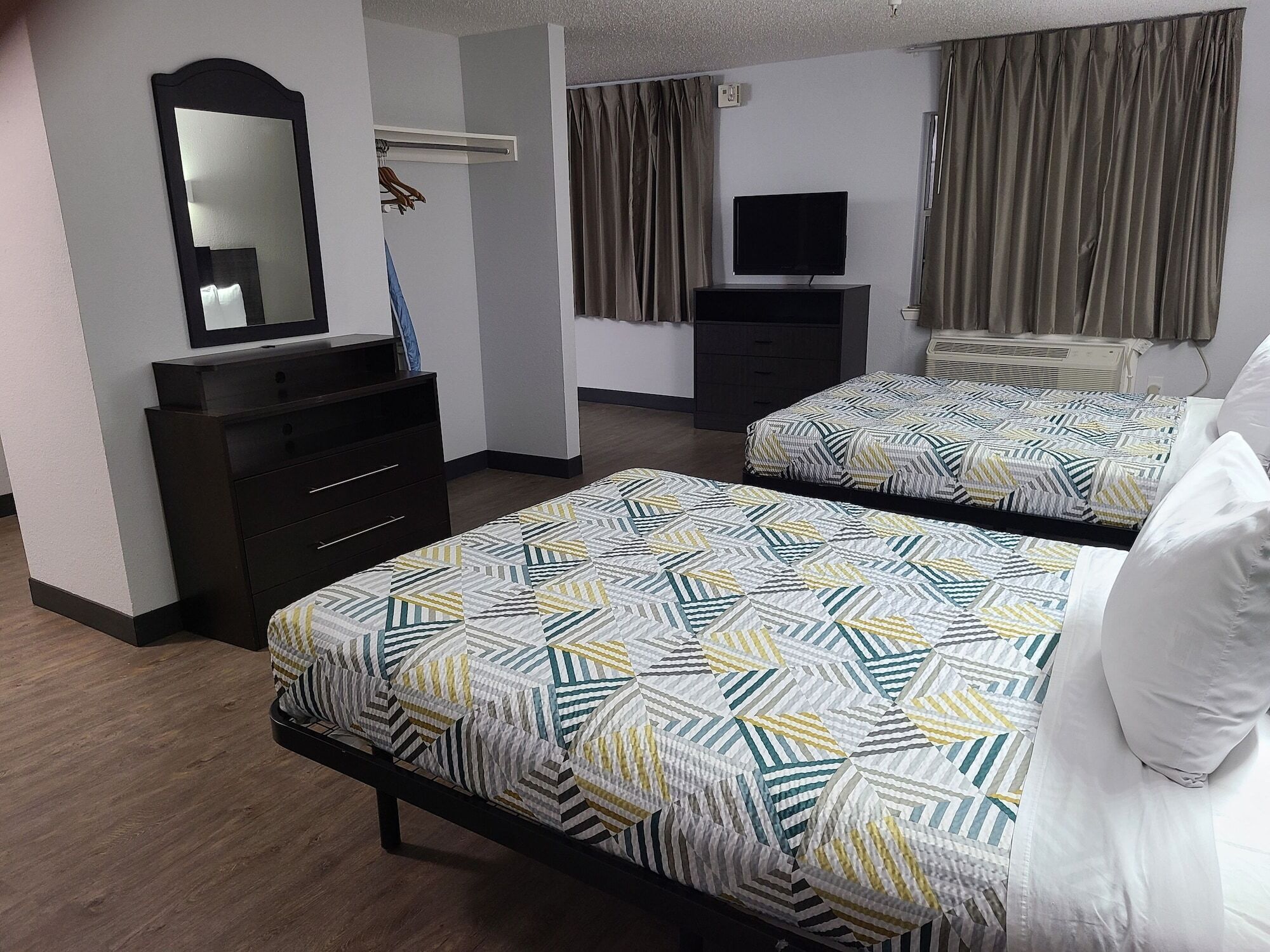 A hotel room with two beds, a dresser, a mirror, a television, and a window with curtains.