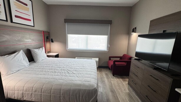 Habitación de hotel con cama, televisión, una silla roja, cómoda y ventana con persianas.