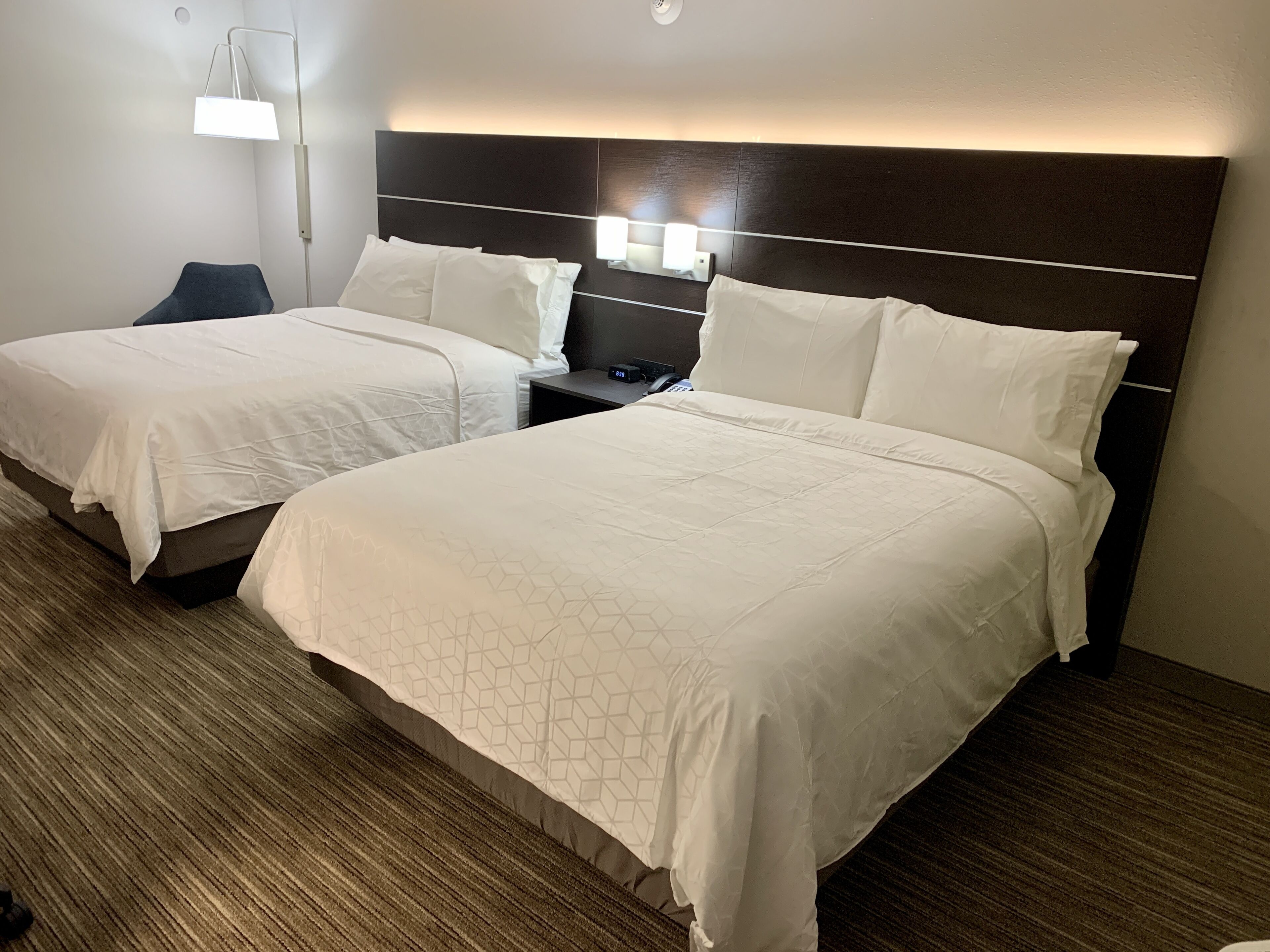 A hotel room with two beds, a nightstand, a lamp, and a wall-mounted light.