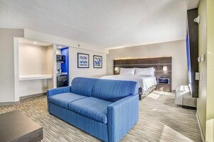 Quarto de hotel com sofá azul, cama com roupa de cama branca, micro-ondas e uma pequena cozinha compacta.