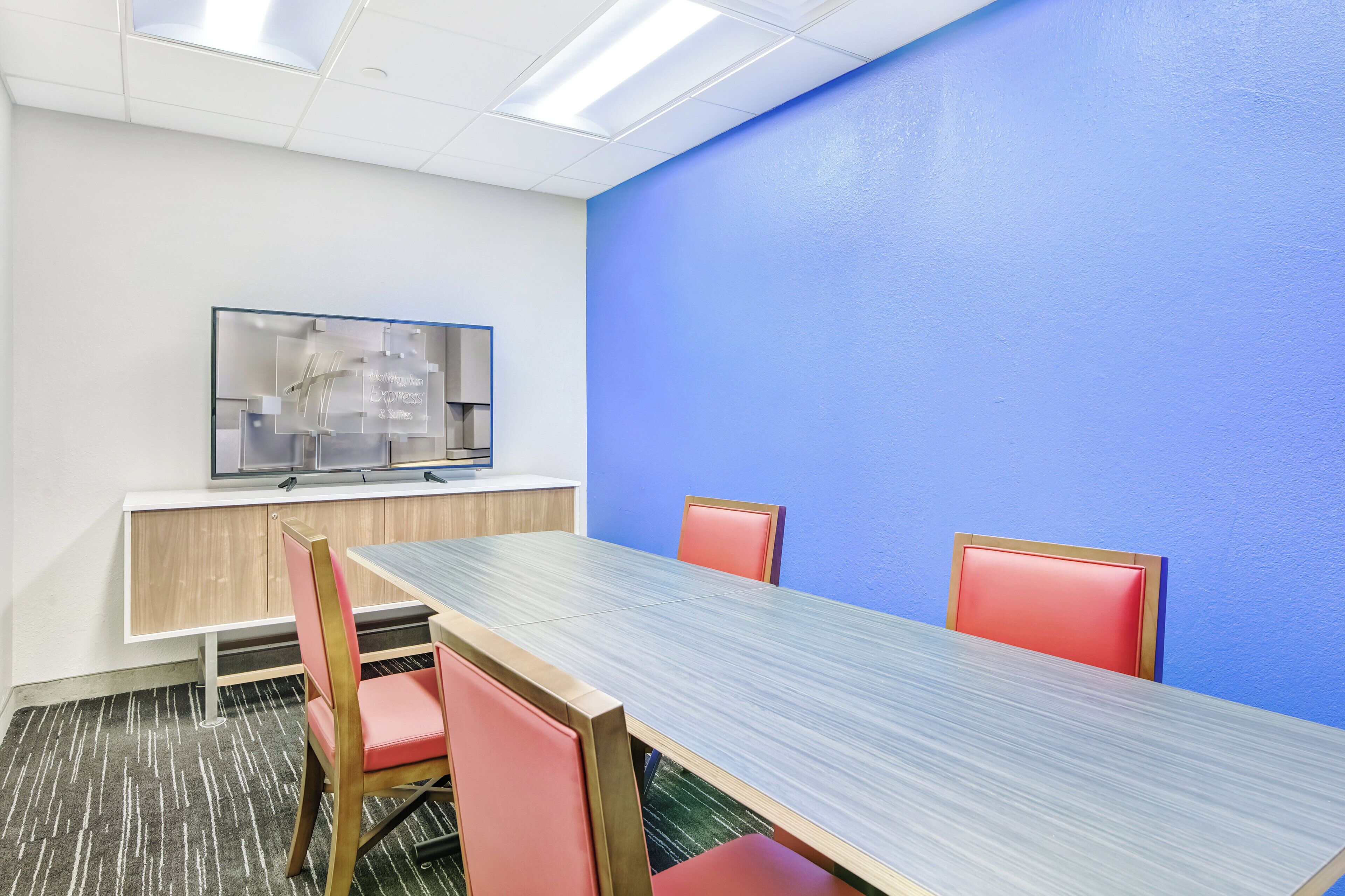 Una sala de conferencias con una mesa larga, dos sillas, un televisor sobre un pedestal y una pared azul decorativa.
