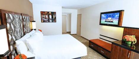 A hotel room with a large bed, a TV mounted on the wall, a desk with a chair, and a small seating area.