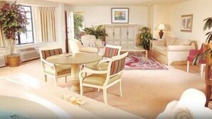 A spacious living room with a round table, a sofa, and a chair. There is a large window with curtains, a cabinet, and a plant.