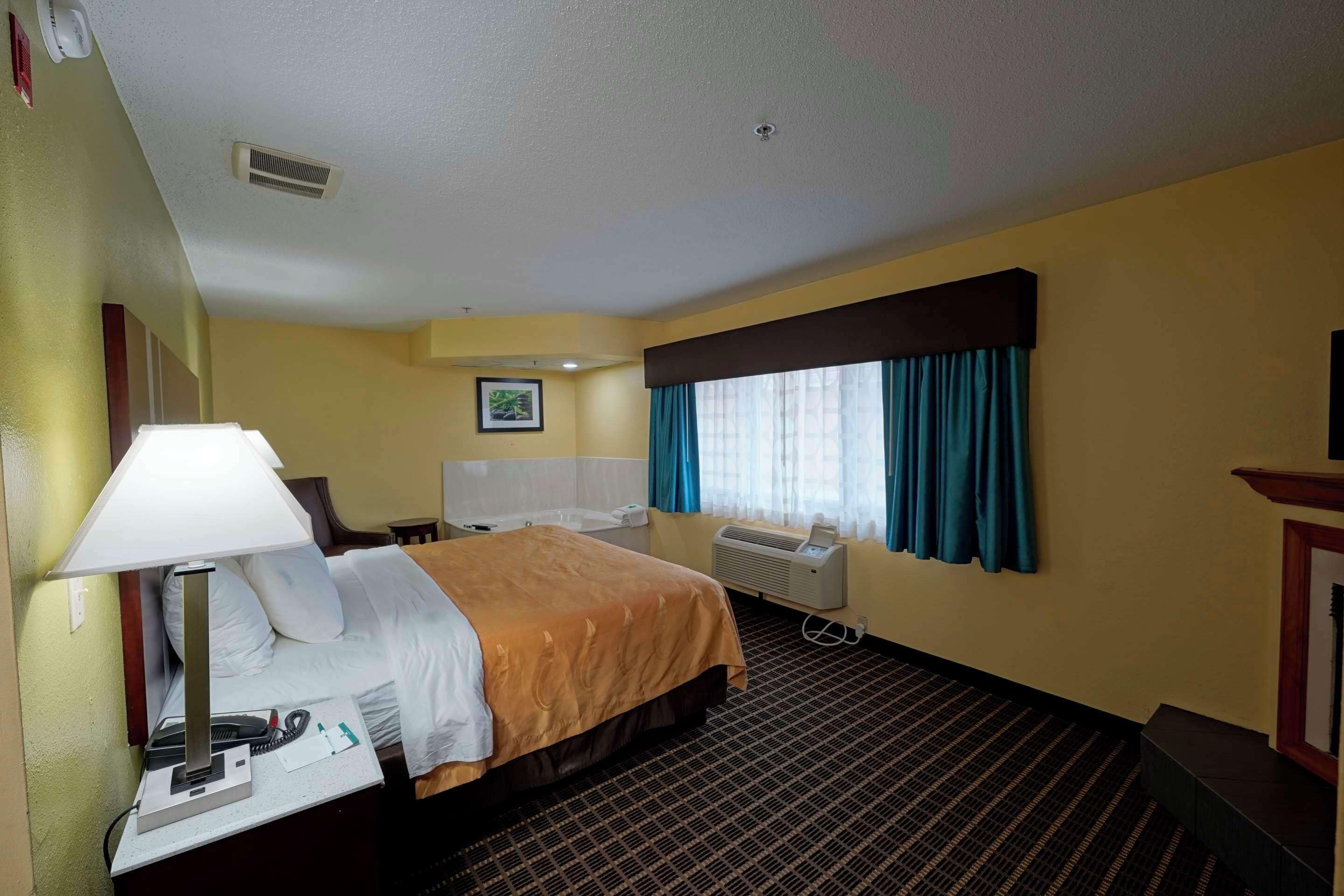 A hotel room with a bed, bedside table with a lamp, a desk with a telephone, a chair, a window with curtains, an air conditioning unit, and a picture on the wall.