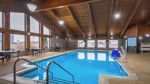 Une piscine intérieure offrant une vue dégagée grâce à de grandes fenêtres, avec des poutres en bois au plafond et un espace détente comprenant des tables et des chaises.