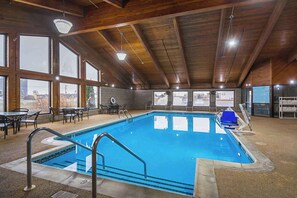 Una piscina interna con ampie finestre che offrono una vista aperta, travi in legno al soffitto e un'area relax con tavoli e sedie.