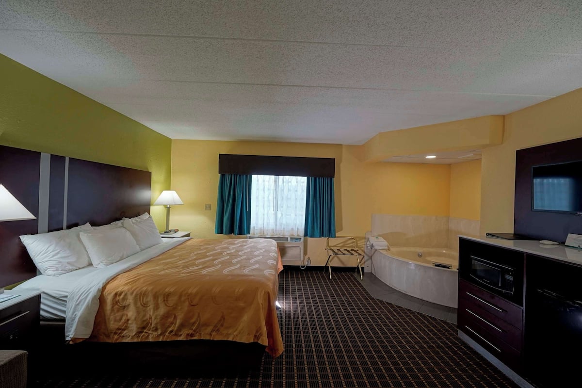 A hotel room with a bed, a desk, a desk lamp, a TV, a bathtub, and a window with curtains.