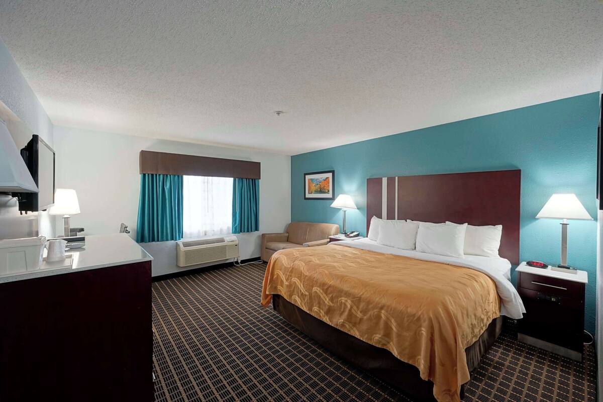 A hotel room with a large bed, a desk with a lamp, a chair, and a window with curtains.