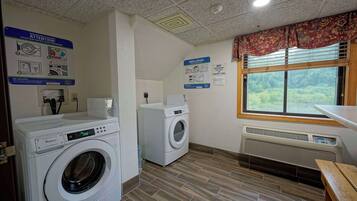 A laundry room with two Whirlpool washers, a wooden bench, a window with a view of greenery, and safety signs on the walls.