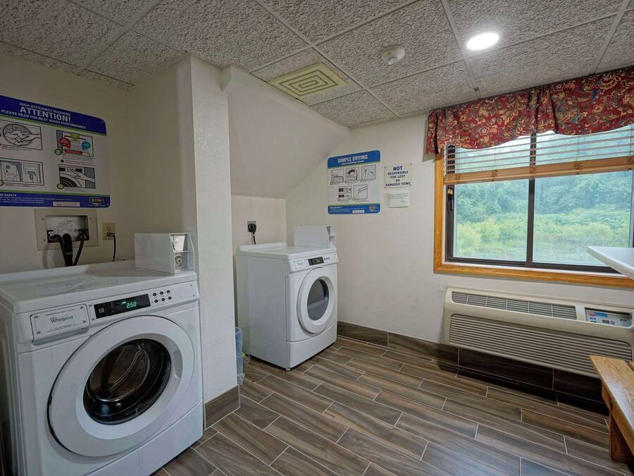 A laundry room with two Whirlpool washers, a wooden bench, a window with a view of greenery, and safety signs on the walls.