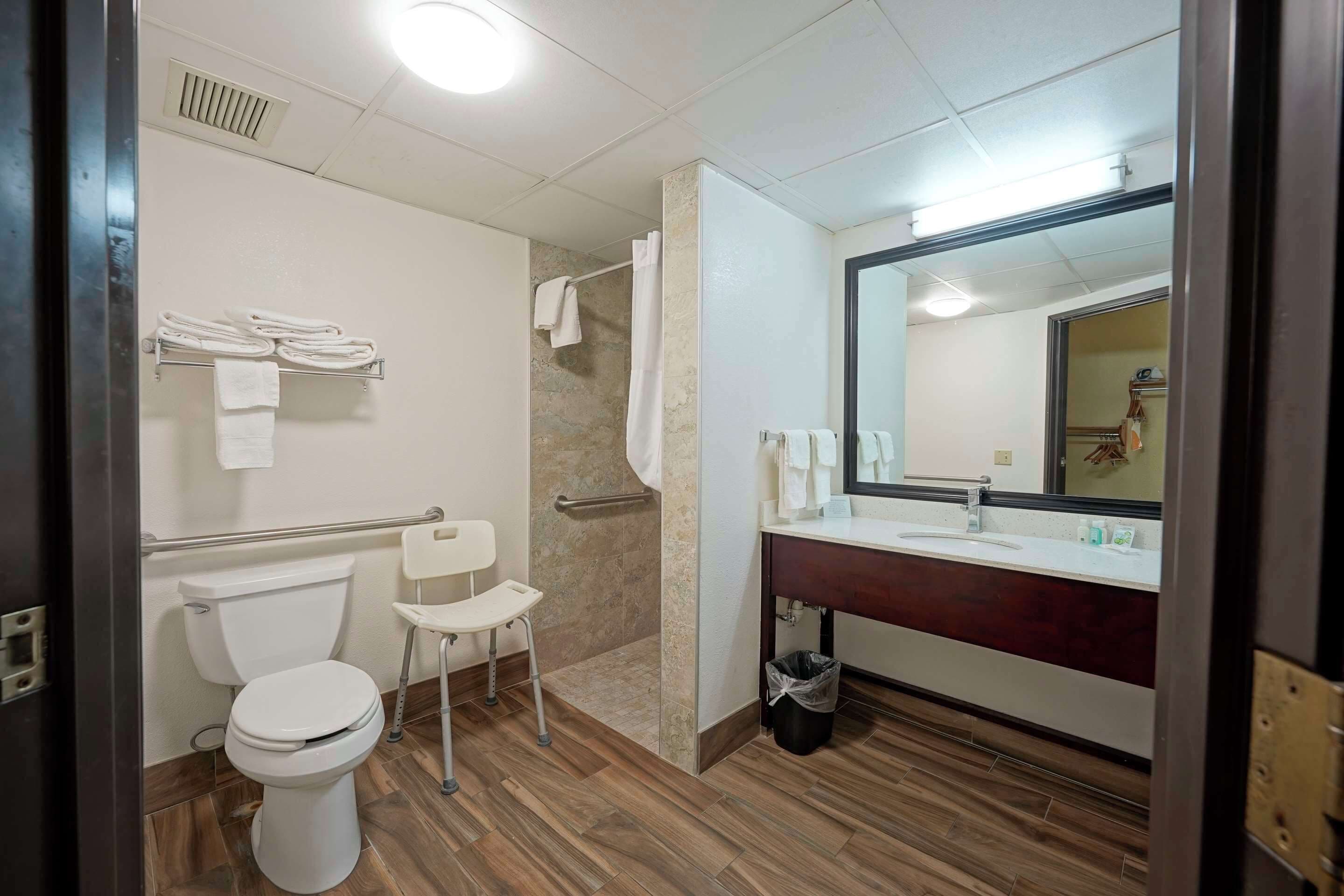 A bathroom with a toilet, a shower with a grab bar, a sink with a mirror, and a wheelchair-accessible chair.