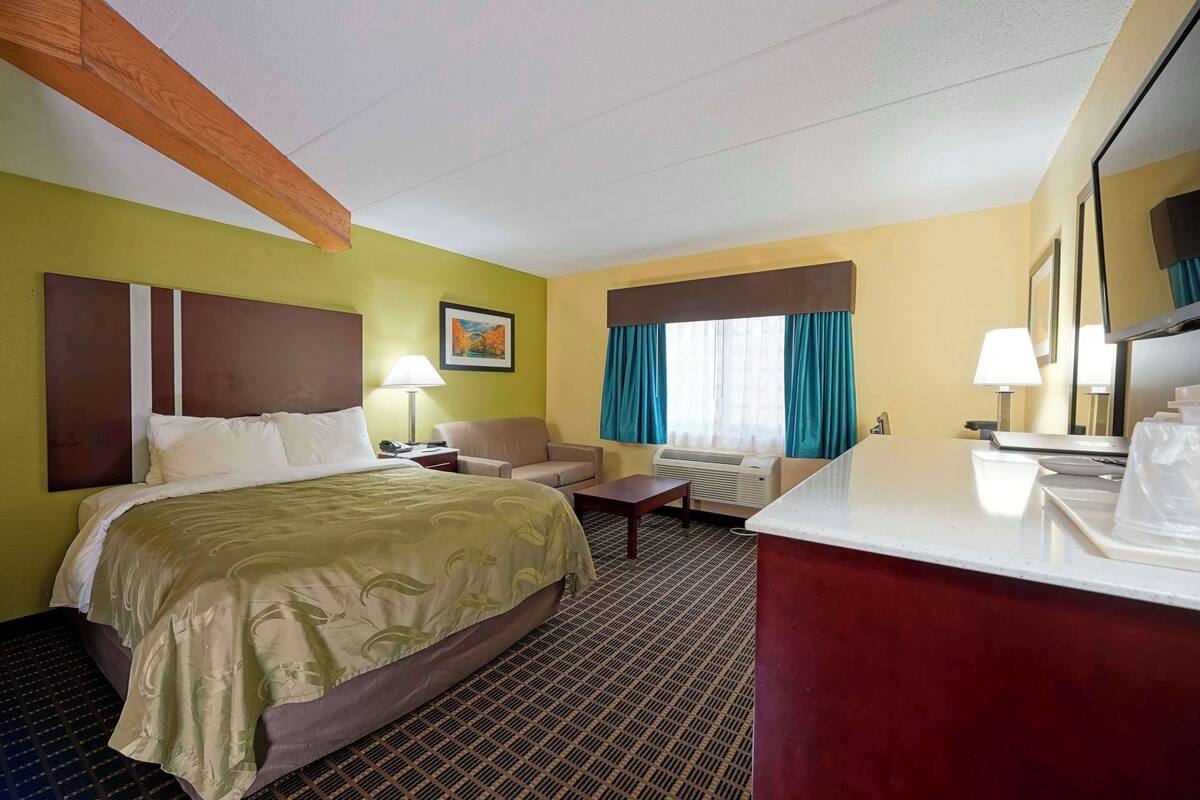 A hotel room with a bed, a desk, a chair, a sofa, a TV, and a window with curtains.