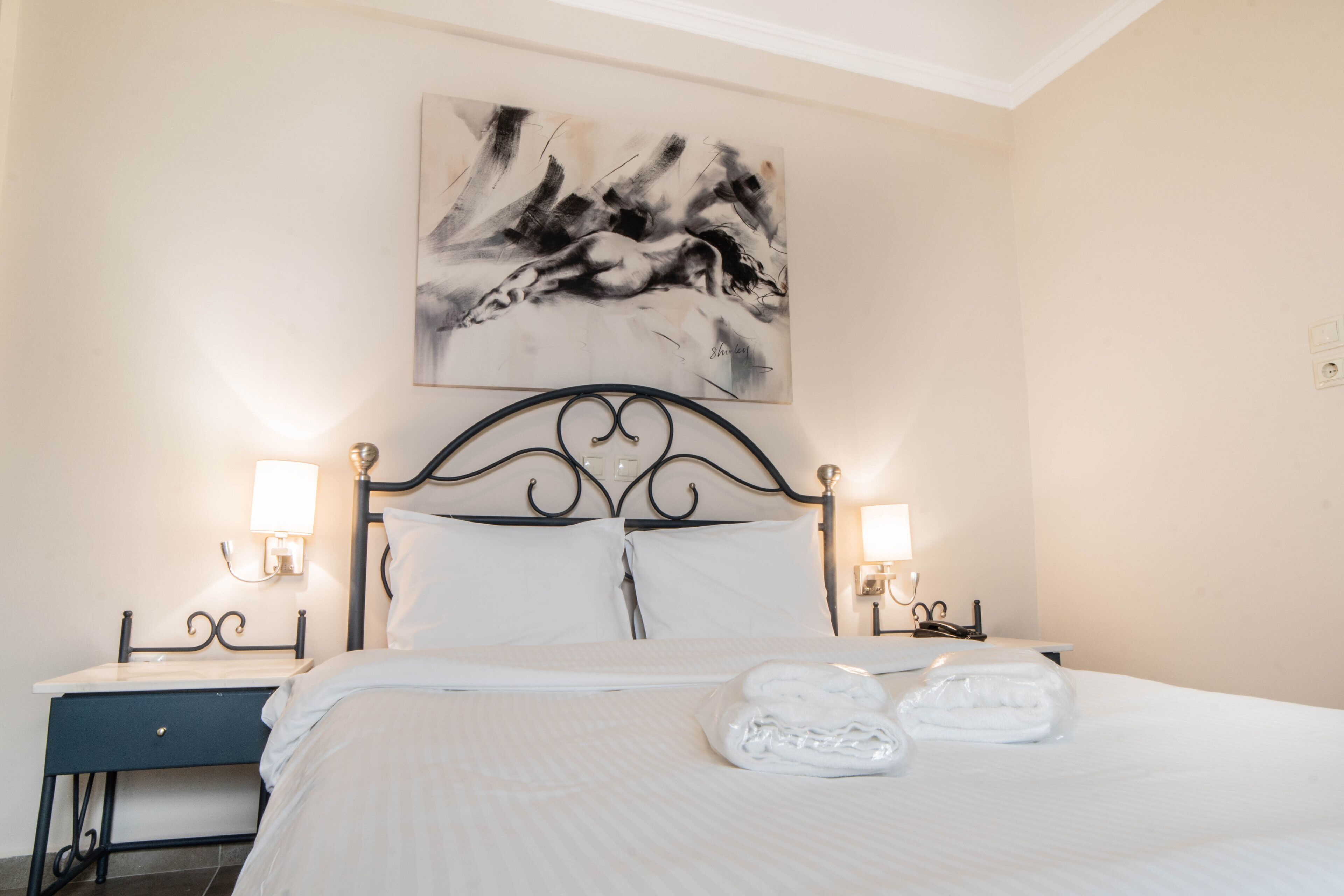 A neatly made bed with white linens and two folded towels, a black metal headboard, bedside lamps, and a wall-mounted abstract painting.