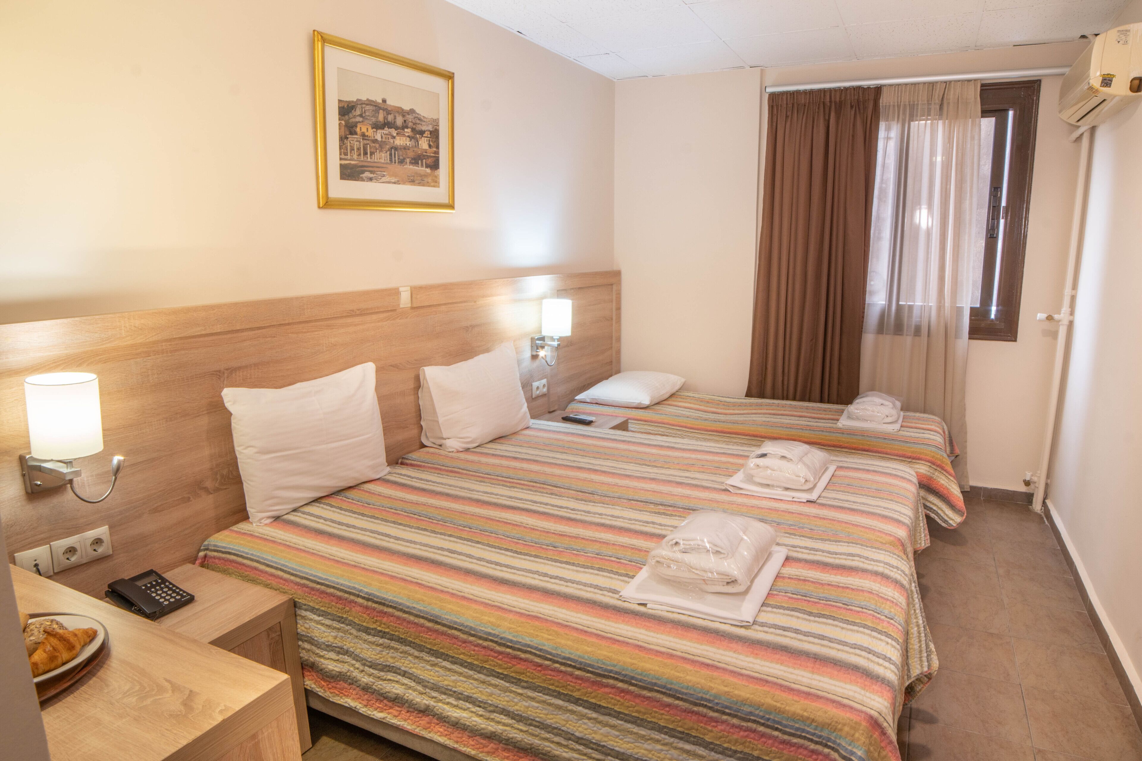 A hotel room with two beds, a bedside table with a lamp, a framed picture on the wall, and a window with curtains.