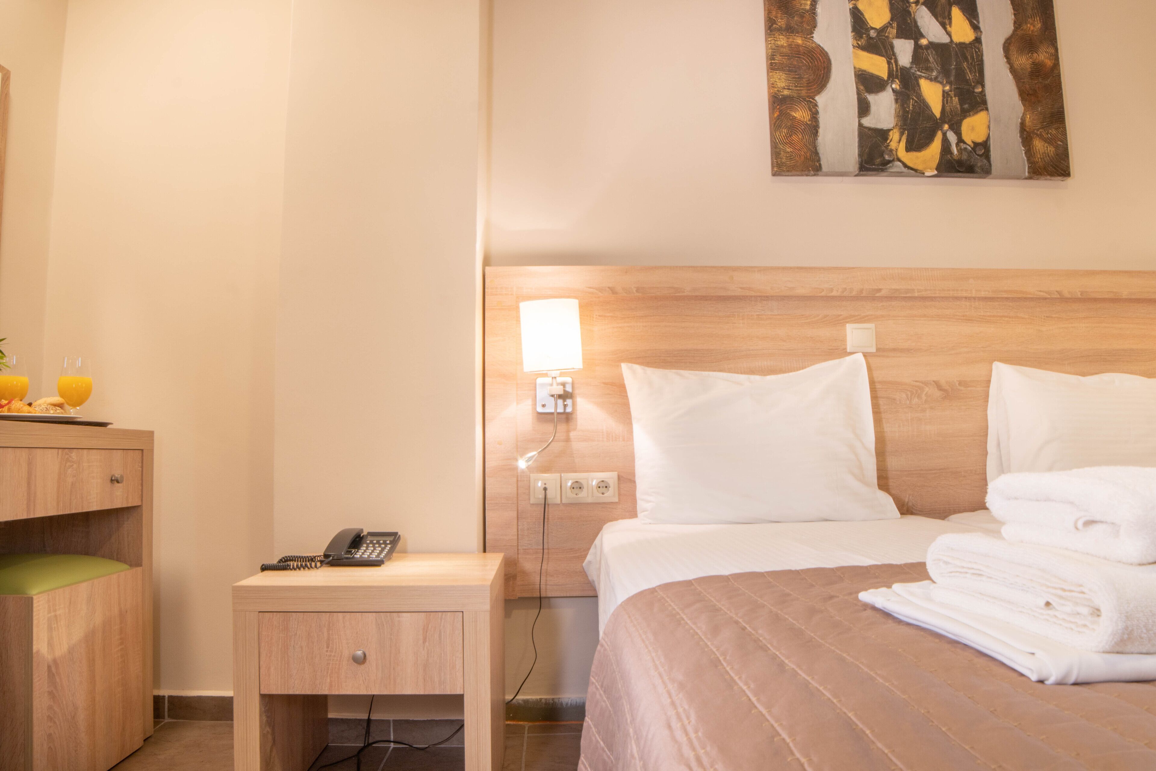 Habitación de hotel con cabecera de madera, mesita de noche con teléfono, luz empotrada en la pared y cama con ropa blanca y toallas dobladas.