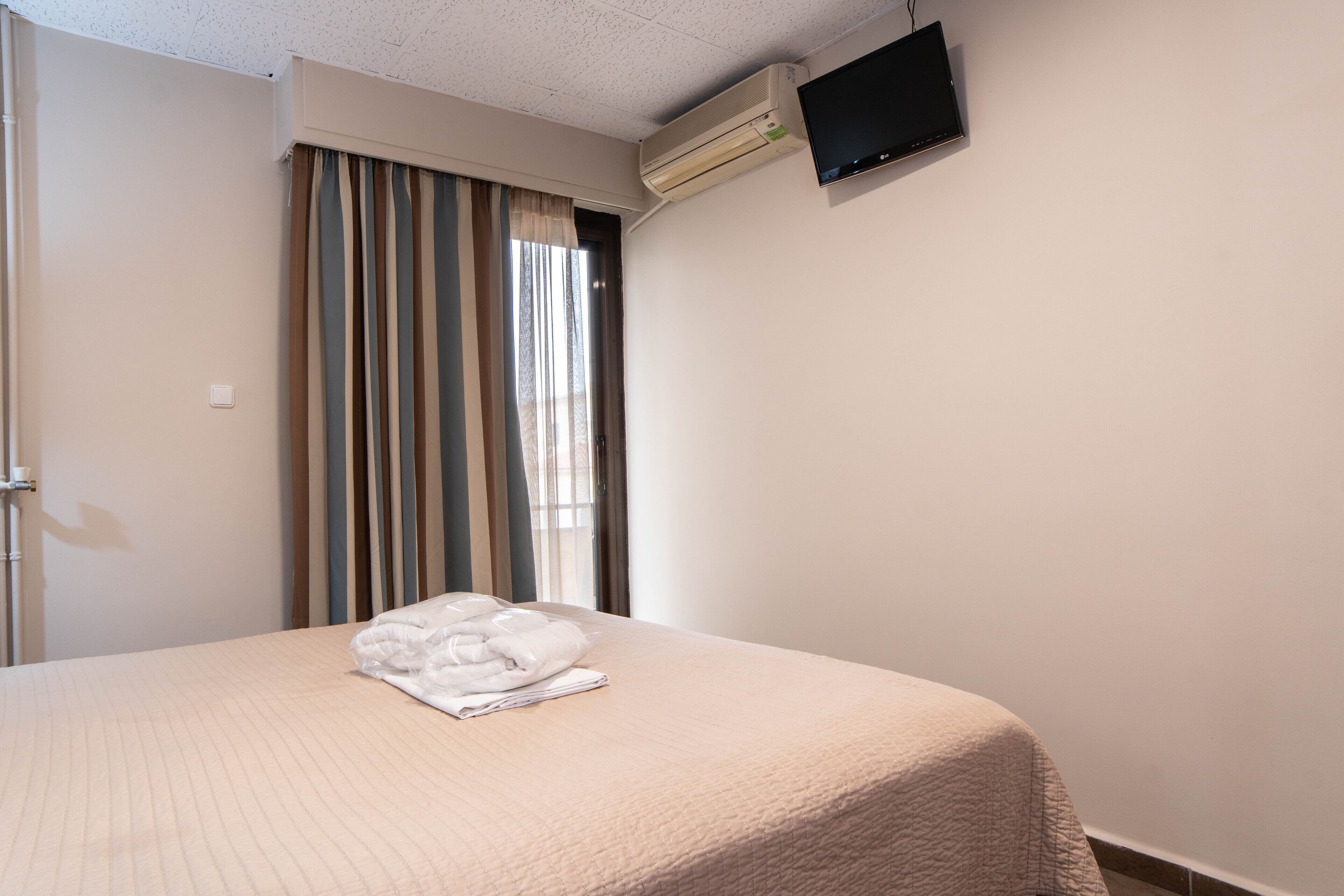 A hotel room with a bed, a flat-screen TV mounted on the wall, and an air conditioning unit.