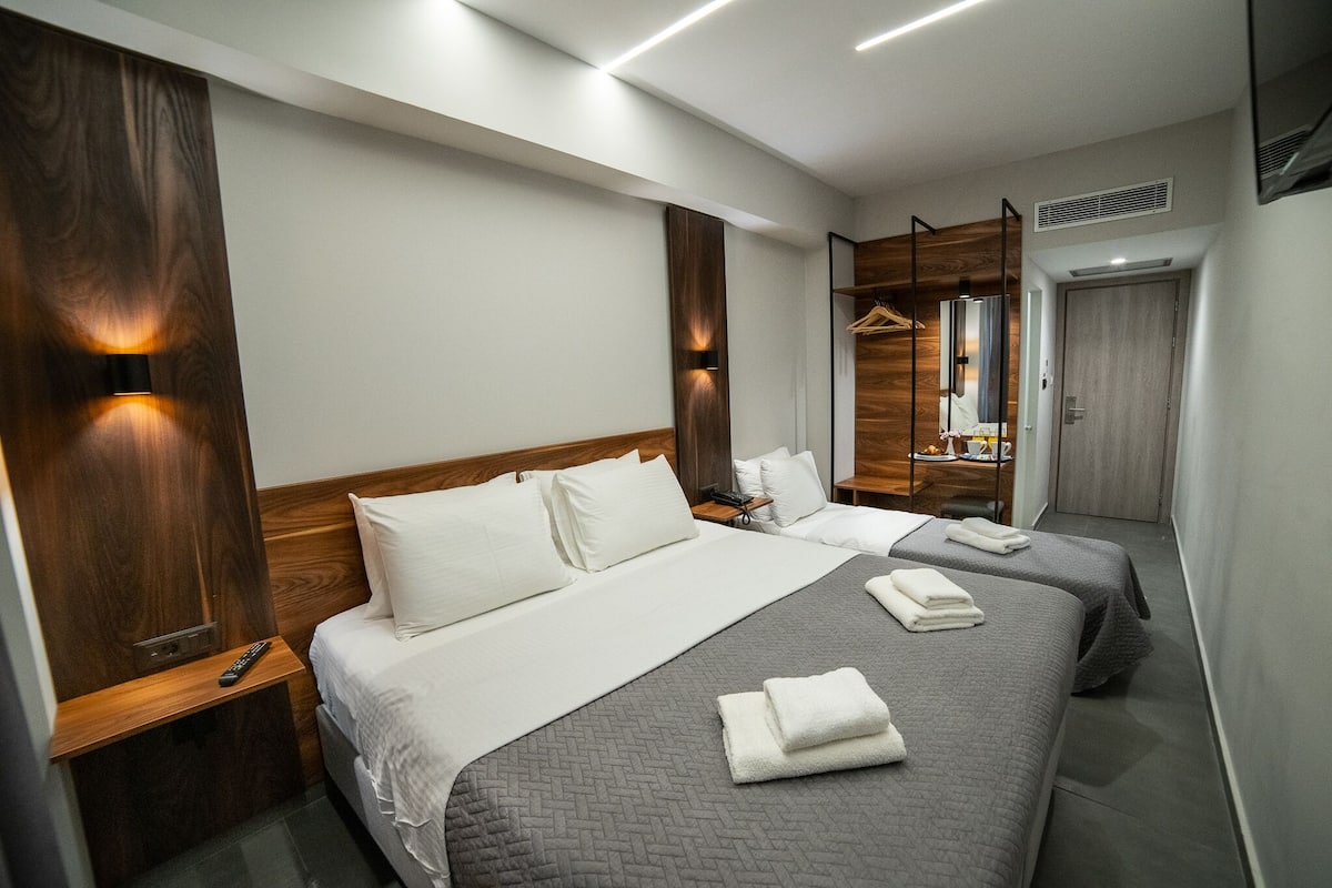 Hotellihuone, jossa on kaksi sänkyä, puinen vaatekaappi, peili ja henkariteline vaatteille.
