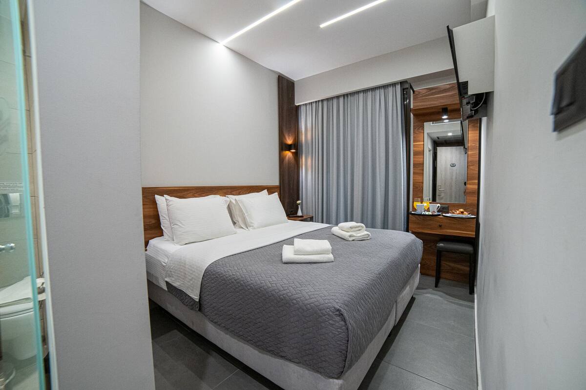 Moderni hotellihuone, jossa on siististi pedattu sänky, puinen pääty, pieni työpöytä peilillä sekä lasiseinän kautta näkyvä kylpyhuone.
