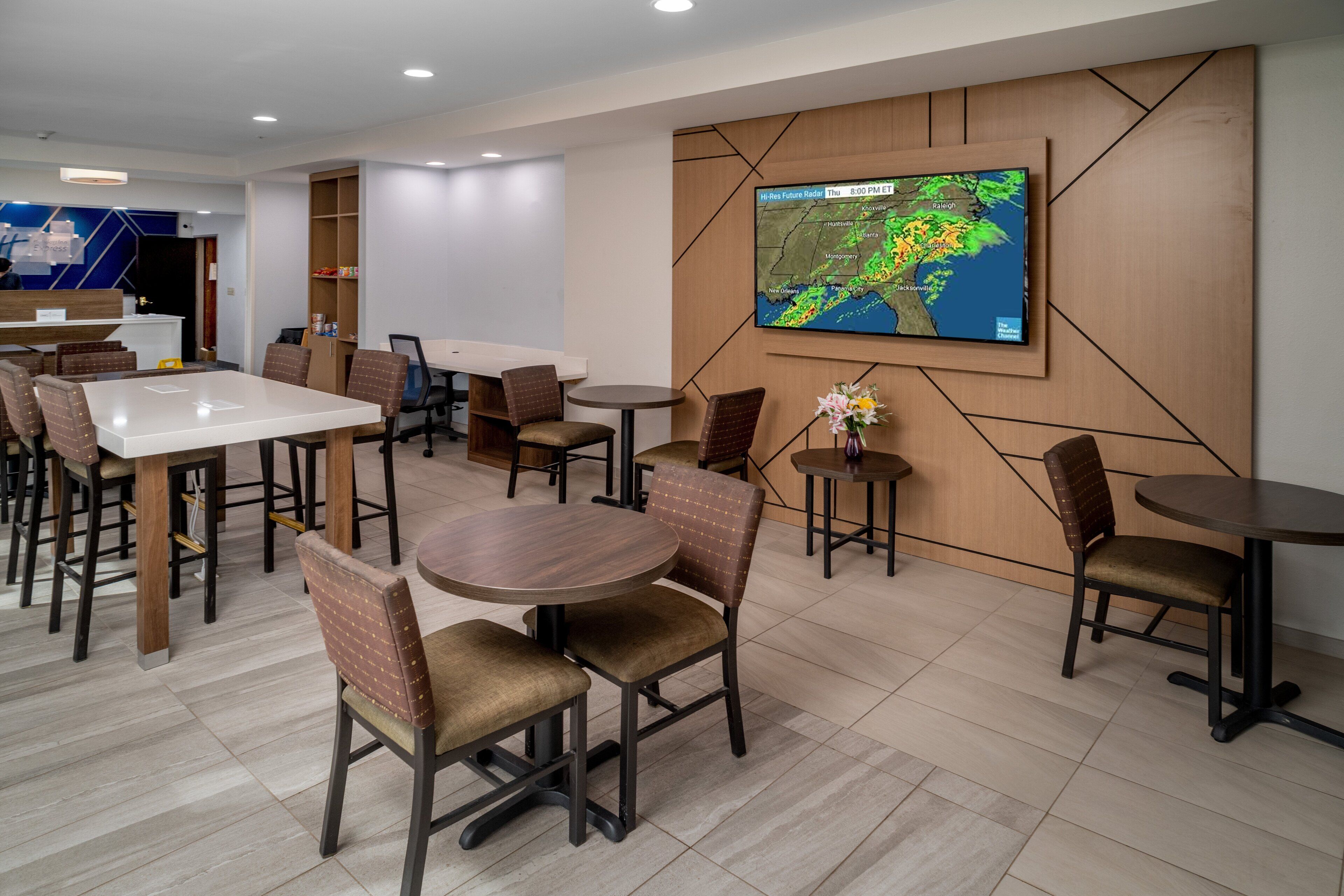 Un moderno vestíbulo de hotel con un televisor grande mostrando un mapa del tiempo, varias zonas de estar con mesas y sillas, y un mostrador de recepción.