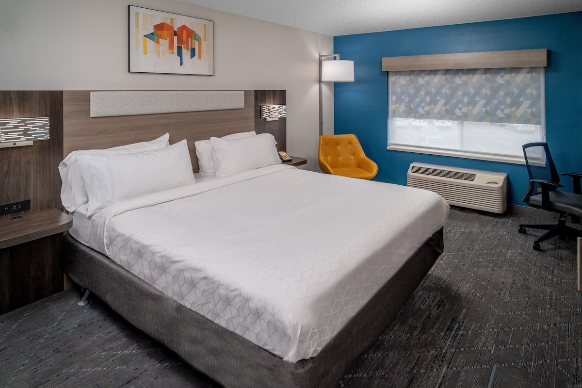 A hotel room with a large bed, a desk, a chair, a window with blinds, and a wall-mounted air conditioner.