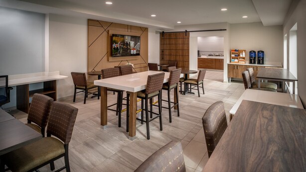 A modern dining area with multiple tables and chairs, a kitchen area with coffee machines, and a television mounted on the wall. - Holiday Inn Express Charleston-Kanawha City by IHG (Charleston)