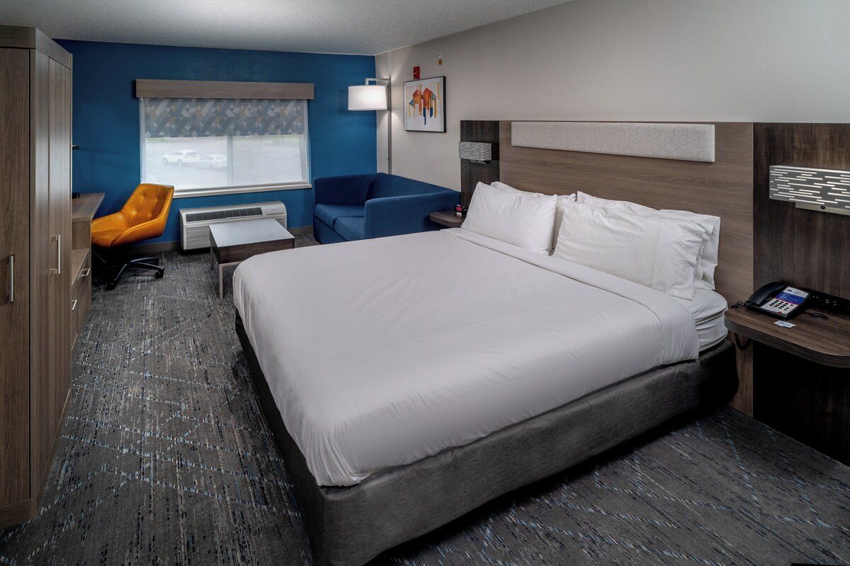 A hotel room with a large bed, a desk, a blue armchair, and a window with a view of parked cars.