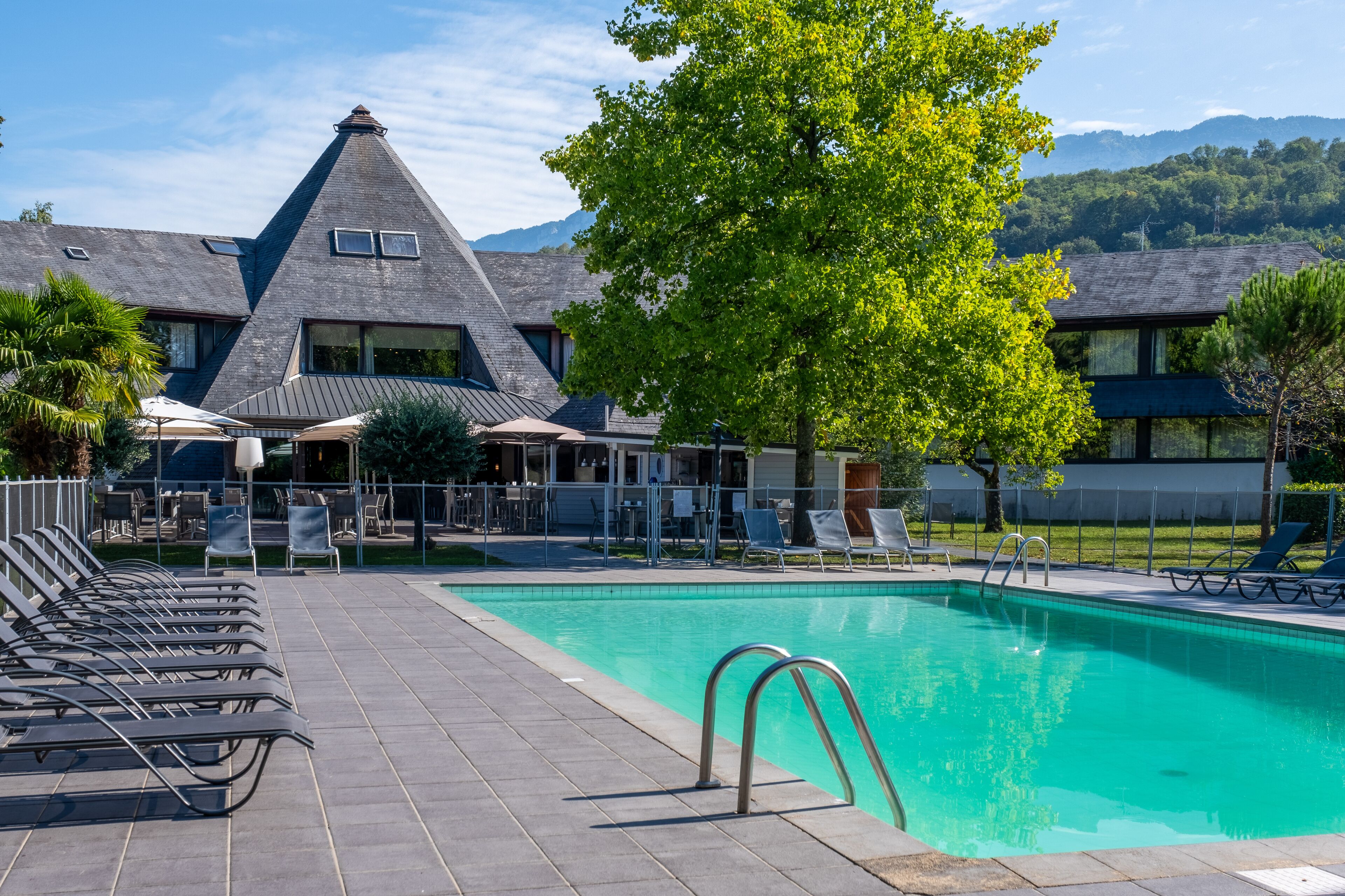 Une piscine avec des chaises longues, un espace de détente couvert et un bâtiment doté d’un toit original.