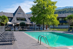 A swimming pool with lounge chairs, a covered seating area, and a building with a distinctive roof.