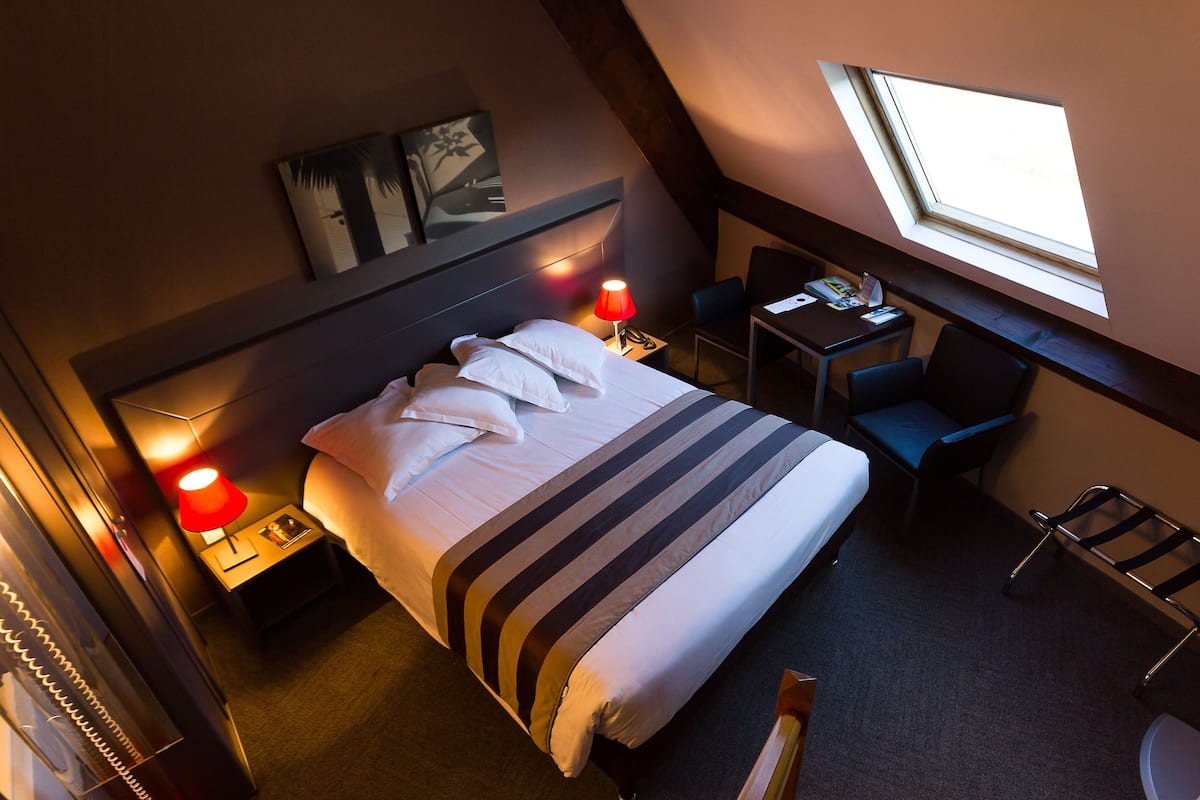 Quarto de hotel com cama, abajures, escrivaninha, cadeira e janela.