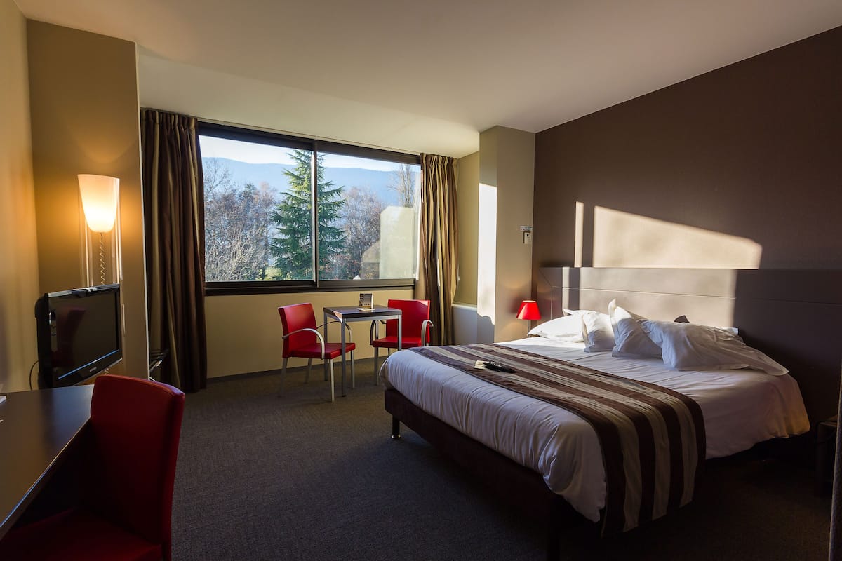 Quarto de hotel com cama, escrivaninha, cadeira, televisão e janela com cortinas.