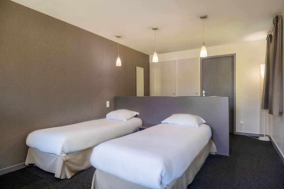 Quarto de hotel com duas camas de solteiro, um espelho grande, um guarda-roupa e um lustre.